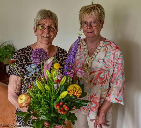 2 vrouwen die naast elkaar staan. Eén houdt een bos bloemen vast
